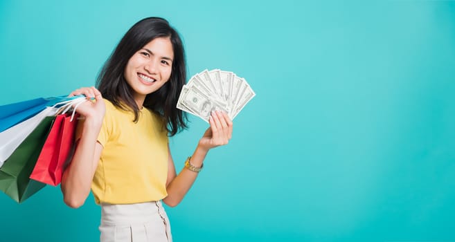 Portrait happy Asian beautiful young woman smile white teeth standing wear yellow t-shirt, She holding shopping bags and dollars money fan, studio shot on blue background with copy space for text