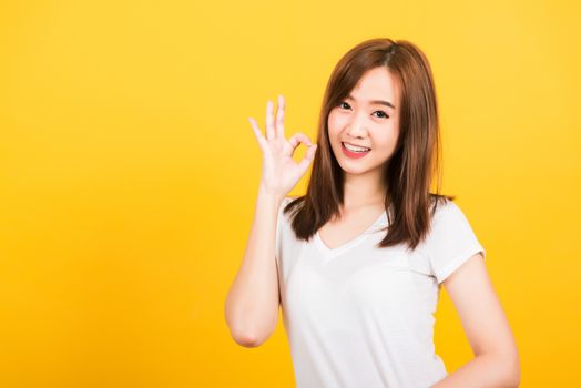 Asian happy portrait beautiful cute young woman teen standing wear t-shirt showing gesturing ok sign with fingers looking to camera isolated, studio shot on yellow background with copy space for text