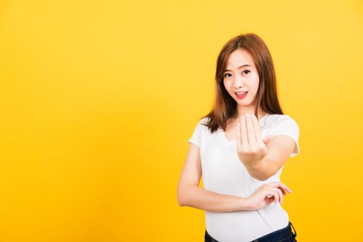 Asian happy portrait beautiful cute young woman teen smile standing wear white t-shirt making gesture hand inviting to come looking to camera isolated, studio shot on yellow background with copy space