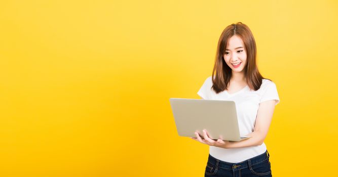 Asian happy portrait beautiful cute young woman teen smiling standing wear t-shirt using laptop computer and showing thumb up looking to camera isolated, studio shot yellow background with copy space