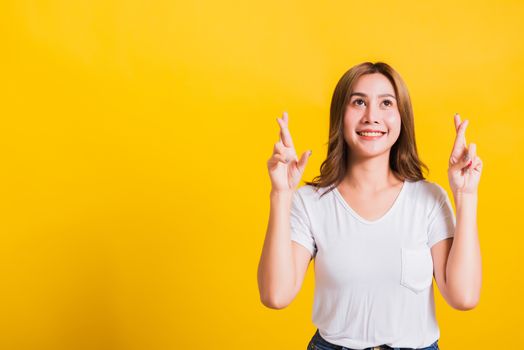 Asian Thai happy portrait beautiful cute young woman smile have superstition her holding fingers crossed for good luck and looking to camera, studio shot isolated on yellow background with copy space