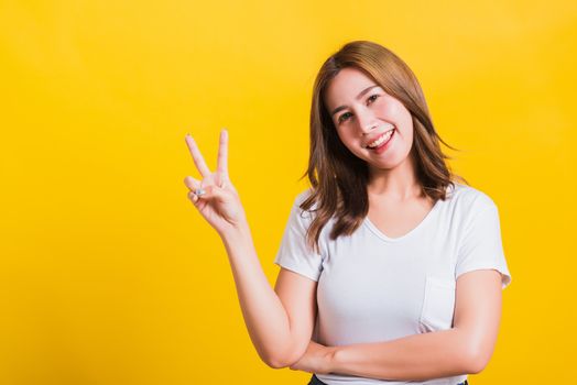 Asian Thai happy portrait beautiful cute young woman smile standing wear t-shirt showing finger making v-sign symbol near eye looking to camera, isolated studio shot yellow background with copy space