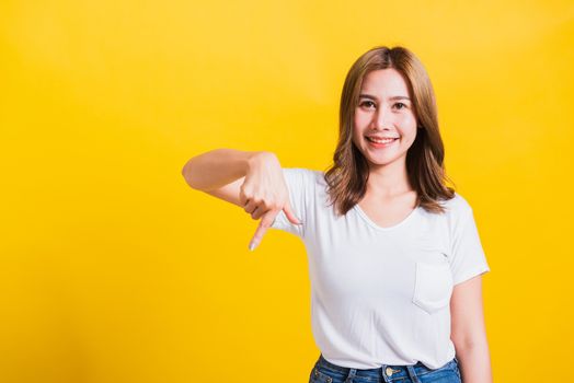 Asian Thai happy portrait beautiful cute young woman standing wear t-shirt makes gesture two fingers point below down and looking to camera, studio shot isolated on yellow background with copy space