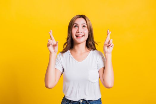 Asian Thai happy portrait beautiful cute young woman smile have superstition her holding fingers crossed for good luck and looking to camera, studio shot isolated on yellow background with copy space