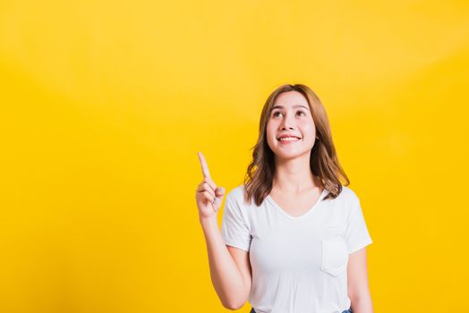 Asian Thai happy portrait beautiful cute young woman standing wear white t-shirt makes gesture two fingers point upwards above looking above, studio shot isolated on yellow background with copy space