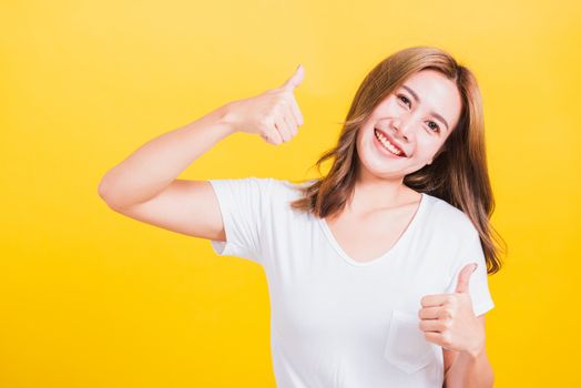 Portrait Asian Thai beautiful happy young woman smiling wear white t-shirt standing successful woman giving two thumbs up gesture sign in studio shot, isolated on yellow background with copy space