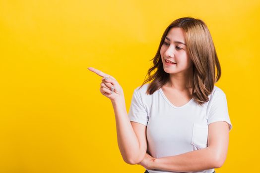 Asian Thai happy portrait beautiful cute young woman standing wear white t-shirt pointing finger away side looking to away side, studio shot isolated on yellow background with copy space