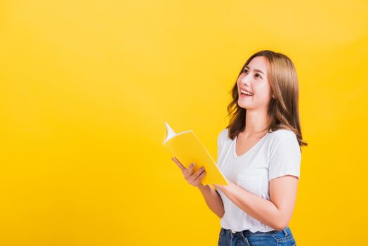 Portrait Asian Thai beautiful happy young lifestyle woman stands holding yellow book or diary she show thumb up finger and looking up away, studio shot isolated on yellow background, with copy space