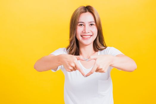 Asian Thai happy portrait beautiful cute young woman smile standing make finger heart figure symbol shape sign with two hands and looking camera, studio shot isolated yellow background with copy space