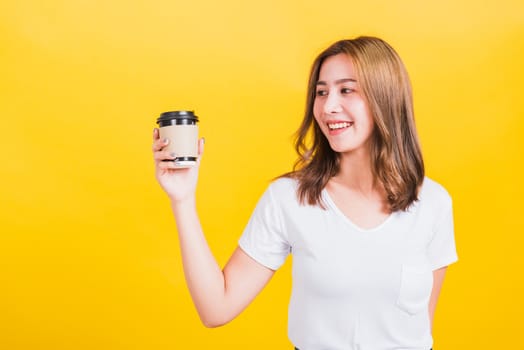 Portrait Asian Thai beautiful happy young woman wear white t-shirt standing smiling holding take away coffee paper cup, studio shot isolated on yellow background, with copy space