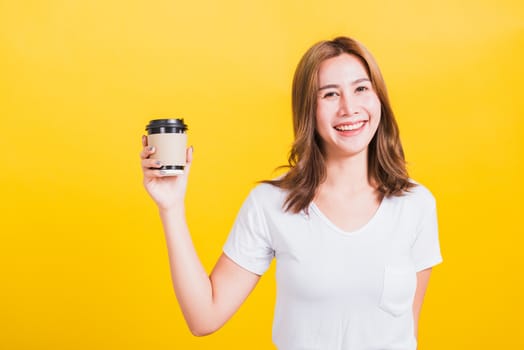 Portrait Asian Thai beautiful happy young woman wear white t-shirt standing smiling holding take away coffee paper cup, studio shot isolated on yellow background, with copy space