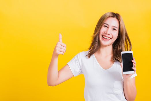 Portrait Asian Thai beautiful happy young woman standing smile, holding blank screen mobile phone and showing thumbs up gesture, shoot photo in studio focus face on yellow background, with copy space