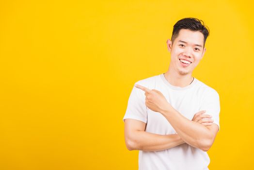 Portrait happy Asian handsome young man smiling standing wearing white t-shirt pointing finger to the side away he looking to camera, studio shot isolated yellow background