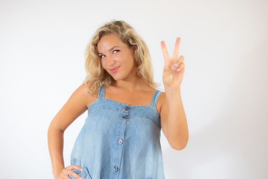 Pretty blonde woman in blue dress smiling making victory gesture