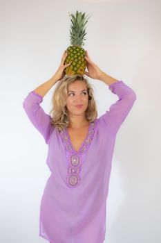 Pretty blonde woman in lila caftan smiling on white background picking up a pineapple on her head