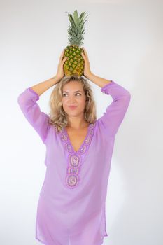 Pretty blonde woman in lila caftan smiling on white background picking up a pineapple on her head