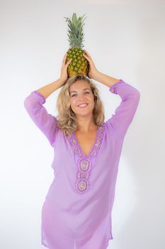 Pretty blonde woman in lila caftan smiling on white background picking up a pineapple on her head