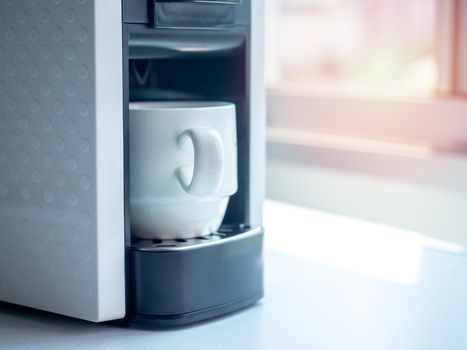 White ceramic coffee cup on coffee machine on white table near the window with sunlight in the morning with copy space.