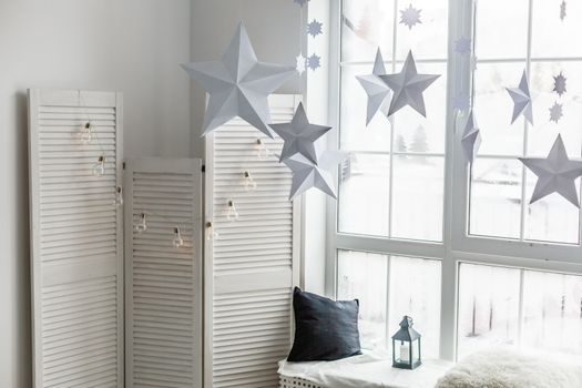 Large bright window. On the window-sill is a gray knitted plaid with gifts and sparkles with herlands. Paper Christmas stars are suspended on the background of the window.