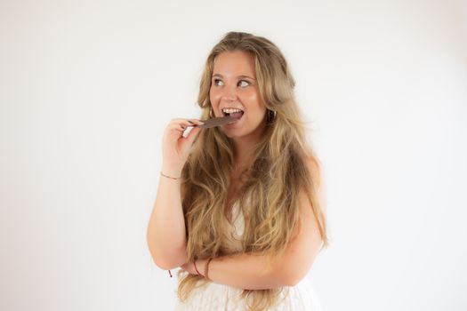 Portrait of a pretty blonde girl in a white dress eating a piece of chocolate