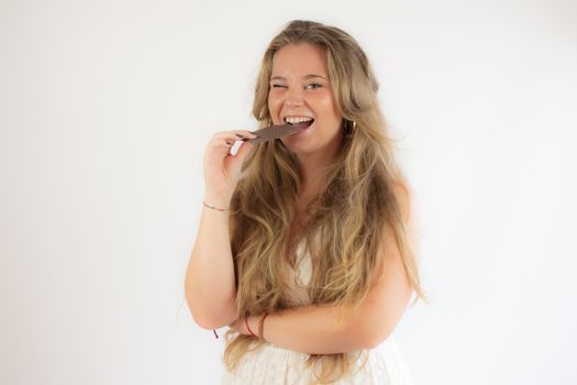 Portrait of a pretty blonde girl in a white dress eating a piece of chocolate