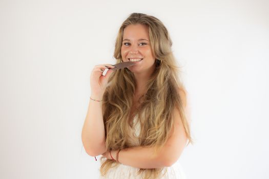 Portrait of a pretty blonde girl in a white dress eating a piece of chocolate