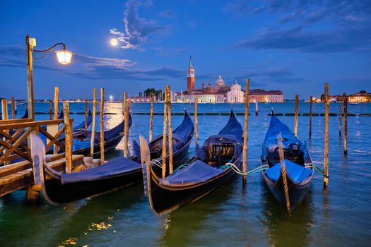 Romantic vacation Venice travel background - gondolas at Saint Mark (San Marco) square and Basilica San Giorgio Maggiore Church seen across Venice lagoon with full moon. Venice, Italy