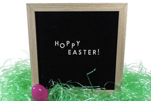 A Black Sign With a Birch Frame That Says Happy Easter in White Letters With a Pink Easter Egg and Green Easter Grass in Front of It on a Pure White Background