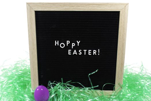 A Black Sign With a Birch Frame That Says Happy Easter in White Letters With a Purple Easter Egg and Green Easter Grass in Front of It on a Pure White Background