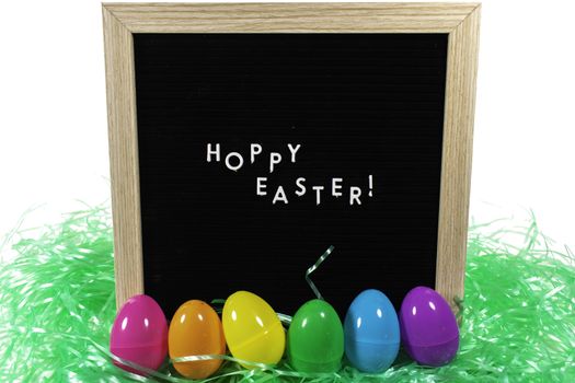 A Black Sign With a Birch Frame That Says Happy Easter in White Letters With a Set of Rainbow Eggs and Green Easter Grass in Front of It on a Pure White Background
