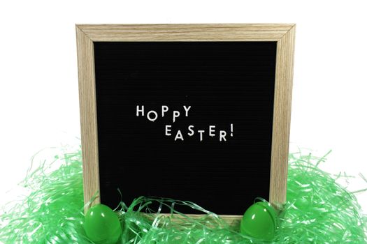 A Black Sign With a Birch Frame That Says Happy Easter in White Letters With Two Green Easter Eggs and Green Easter Grass in Front of It on a Pure White Background