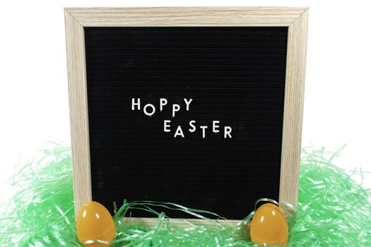 A Black Sign With a Birch Frame That Says Happy Easter in White Letters With Two Orange Easter Eggs and Green Easter Grass in Front of It on a Pure White Background