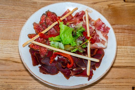 Plate with delicacies. Trio of dried delicacies. Basturma, prosciutto, sausage flat. White plate. Wooden table