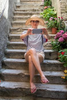 Beautiful female tourist wearing big straw sun hat and shorts sitting and relaxing on old stone house stairs during summer travel on Mediterranean cost on hot summer day and taking selfie.
