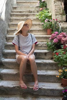 Beautiful female tourist wearing big straw sun hat and shorts sitting and relaxing on old stone house stairs during summer travel Mediterranean costal village on hot summer day.