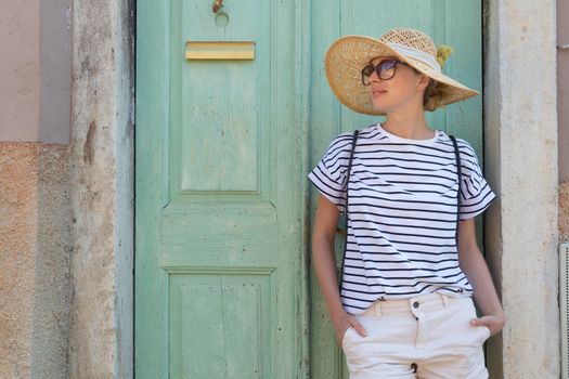 Beautiful young female tourist woman wearing sun hat, standing and relaxing in shade in front of turquoise vinatage wooden door in old Mediterranean town while sightseeing on hot summer day.