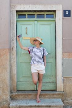 Beautiful young female tourist woman wearing big straw hat, taking self portrait selfie, standing in front of turquoise vinatage wooden door and textured stone wall at old Mediterranean town.