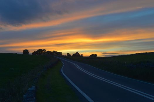 Deep Blue fresh tarmac road in the peak district leads to some trees and a strong orange sunset.