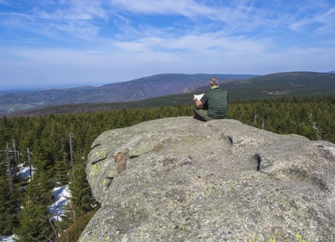 Senior man sitting on rock and holding map on top of holubnik mountain, view of Jizera Mountains jizerske hory with lush green spruce forest, trees, hills and fields, springtime with snow remains, blue sky background. Travell and hiking concept.