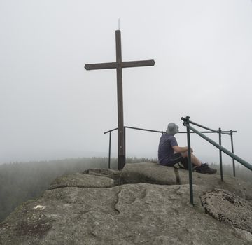young hiker man sitting on the top peak of Jizera mountain rock with big cross in moody day, fog tree background