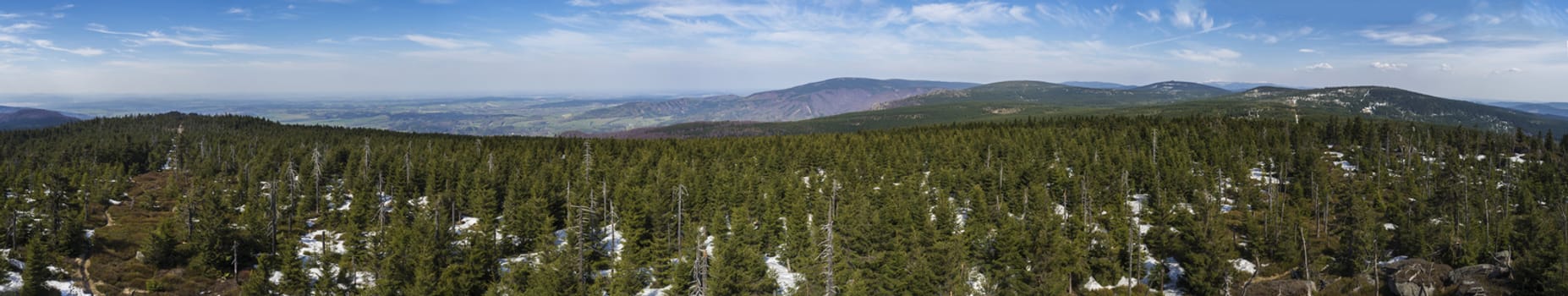 wide panoramic landscape of Jizera Mountains jizerske hory, view from peak of holubnik mountain with lush green spruce forest, trees, hills and fields springtime with snow remains, blue sky background.