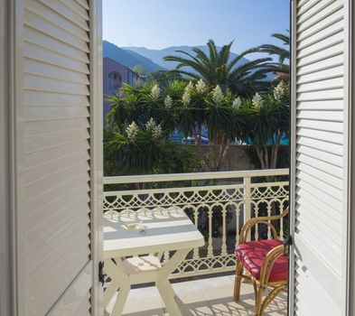 View from hotel room balcony door over white table and red bamboo chair and flowering bush and palm trees on background with pool and mountain at sunny day, Korfu, Greece.