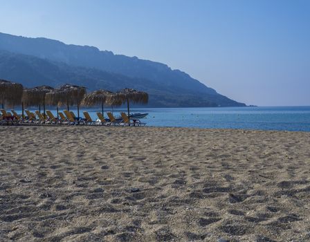 Sand and pebbles beach and turquoise sea shore at Agios Georgios Pagon at Corfu island, Greece with sun umbrellas orange sunbeds and boat, green hill and clear blue sky backgound.