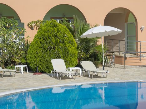Empty hotel swimming pool during sunny day, summer vacation. View of blue water in pool, white sunbeds, umbrella and green palms, Greece, Corfu island.