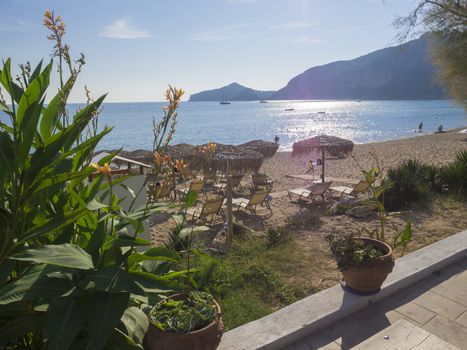 Agios Georgios Pagon sand beach onCorfu island, Greece viewed from promenade with flower pots, with sun beds, swimming peoples and umbrella and view on porto timony bay, claer blue sky backgound.