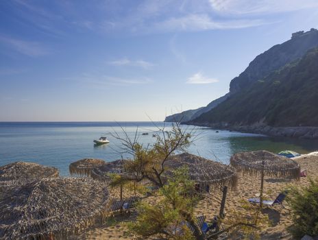 Sand and pebbles beach and turquoise sea shore at Agios Georgios Pagon at Corfu island, Greece with sunbathing peoples and umbrella with view on porto timony bay, claer blue sky backgound.
