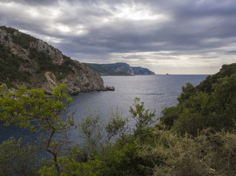View on cllifs, trees and green hill at Paleokastritsa bay, summer cloudy sky, Corfu, Kerkyra, Greece.