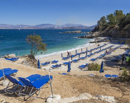 View of Bataria white sand beach with blue sunbeds and tourist people at Kassiopi Bay, and Albanian mountains from the path in Kassiopi, Corfu Island, Greece.