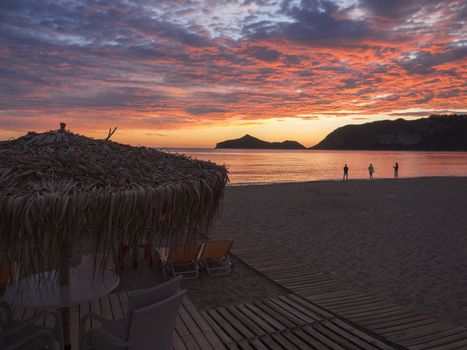 beautiful blue pink orange clouds after sunset on Agios Georgios Pagon beach at Corfu island, Greece with sun umbrellas empty sunbeds and sillhouttes of people admiring the sky, view on porto timony bay.