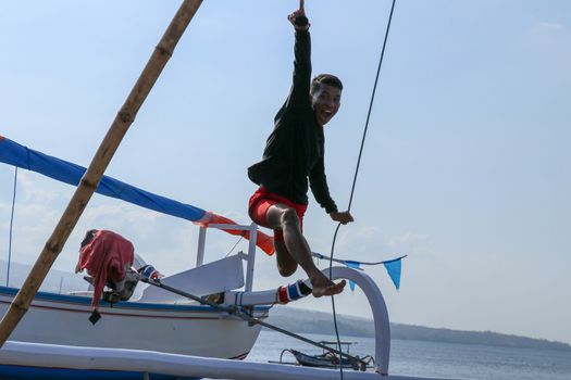 Happy young man jumping on the beach. Young cheerful Indonesian jumps. A teenager in a good mood jumps in the air. In the background a fishing boat. Sunny tropical day with blue sky.